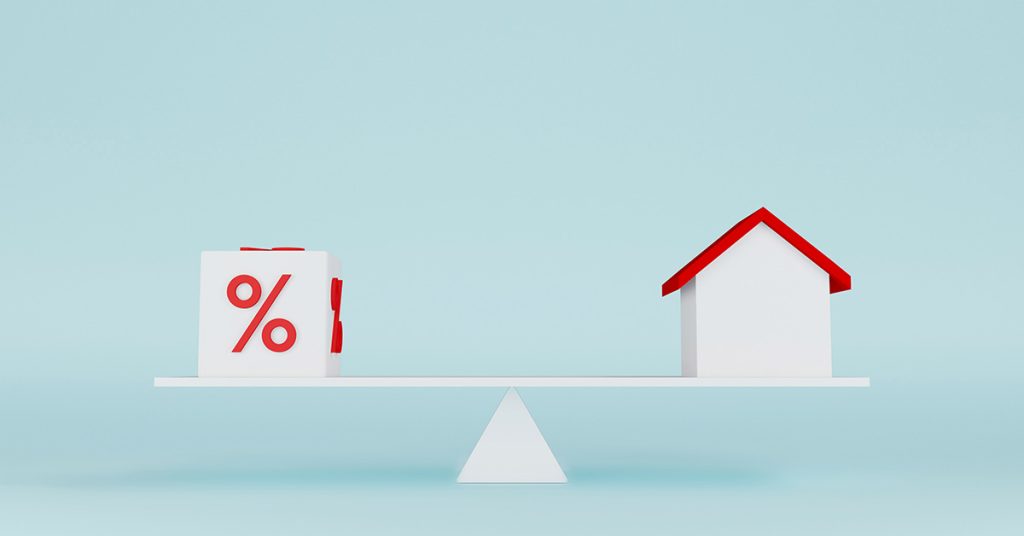 Vor einem hellblauen Hintergrund steht eine Waage, die ein Haus gegen Zinsen abwiegt | Jetzt Immobilie kaufen?